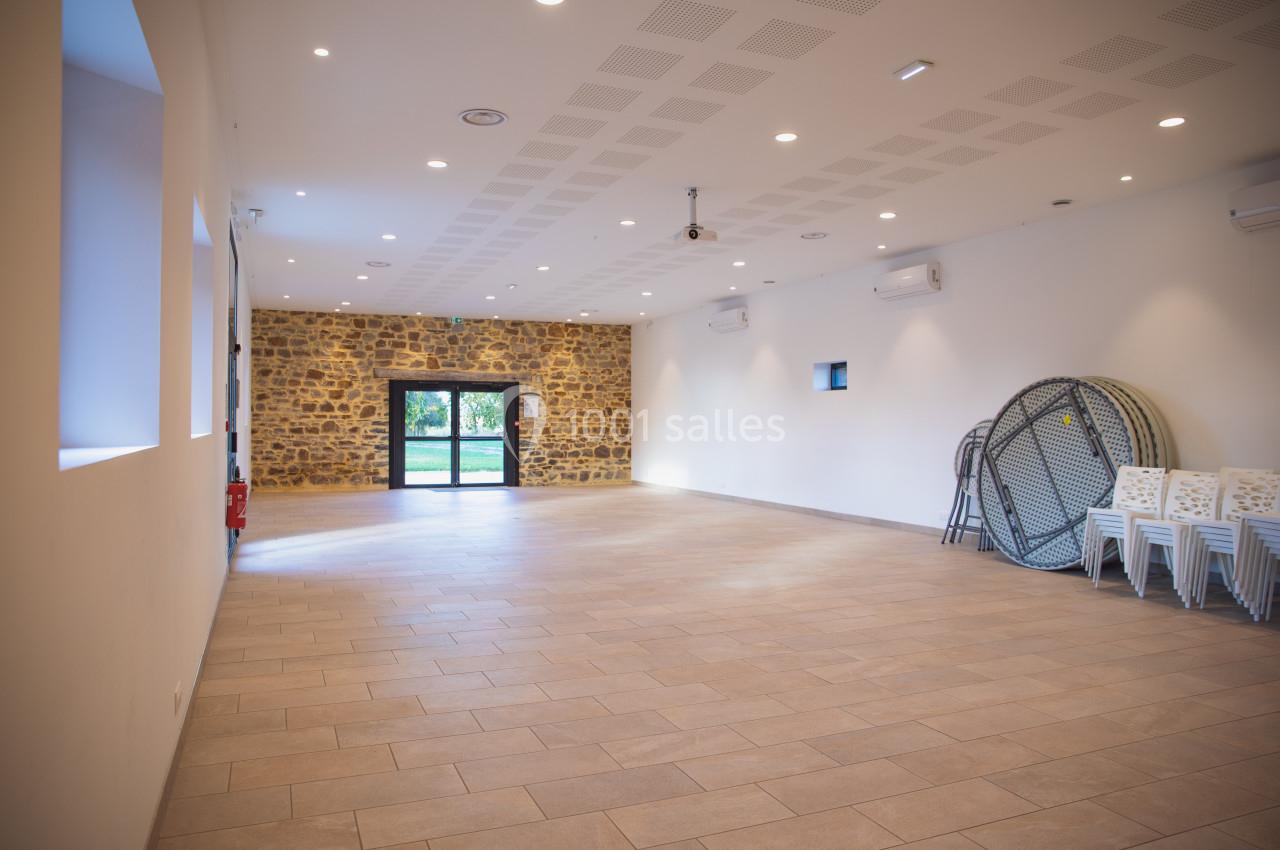 Location salle Domagné (Ille-et-Vilaine) - Domaine de Neuville #12 Salle vide avec murs blancs, mur en pierre au fond, sol carrelé, chaises empilées et tables pliées sur le côté.