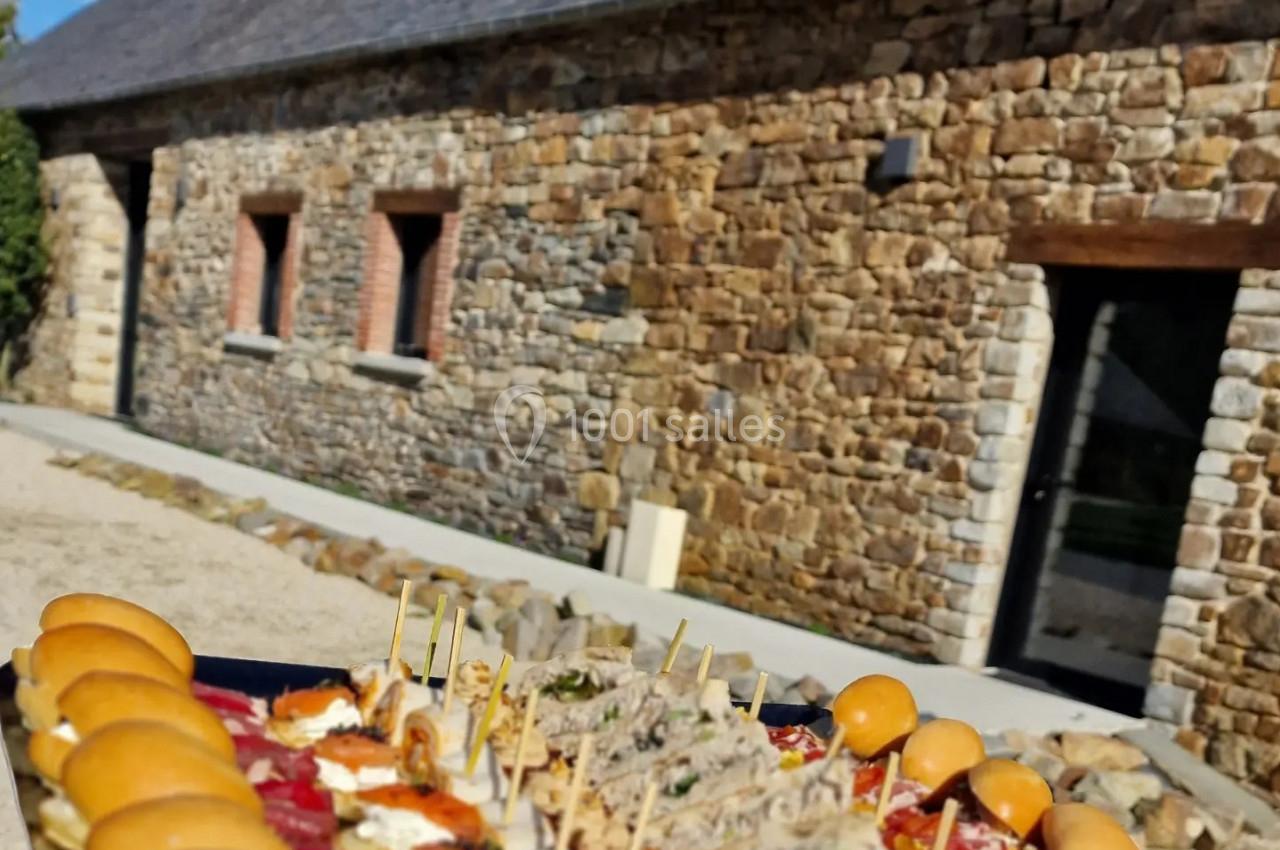 Location salle Domagné (Ille-et-Vilaine) - Domaine de Neuville #17 Plateau de bouchées apéritives variées devant un bâtiment en pierre avec toit en ardoise, sous un ciel bleu.