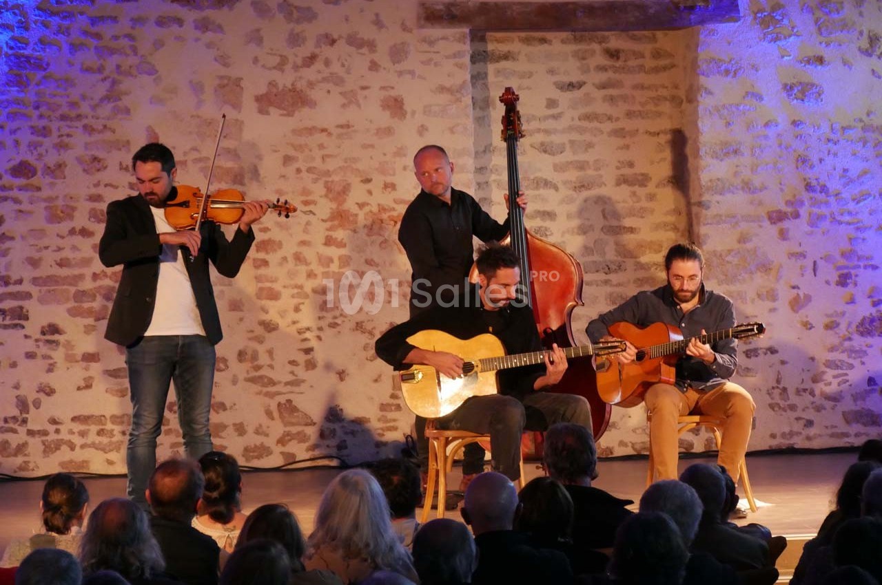 Quatuor de musiciens jouant du violon, de la contrebasse et des guitares devant un public dans une salle en pierre.