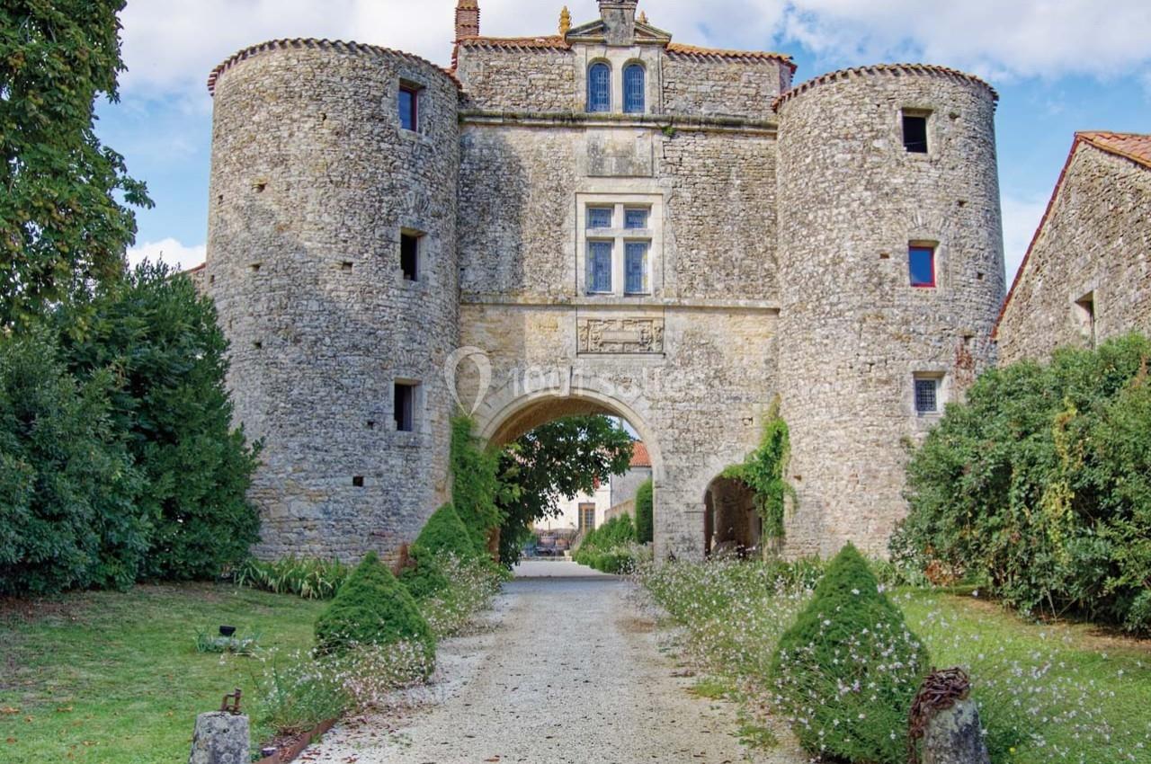Entrée d'un château en pierre avec deux tours rondes, entourée de verdure et d'un chemin gravillonné.
