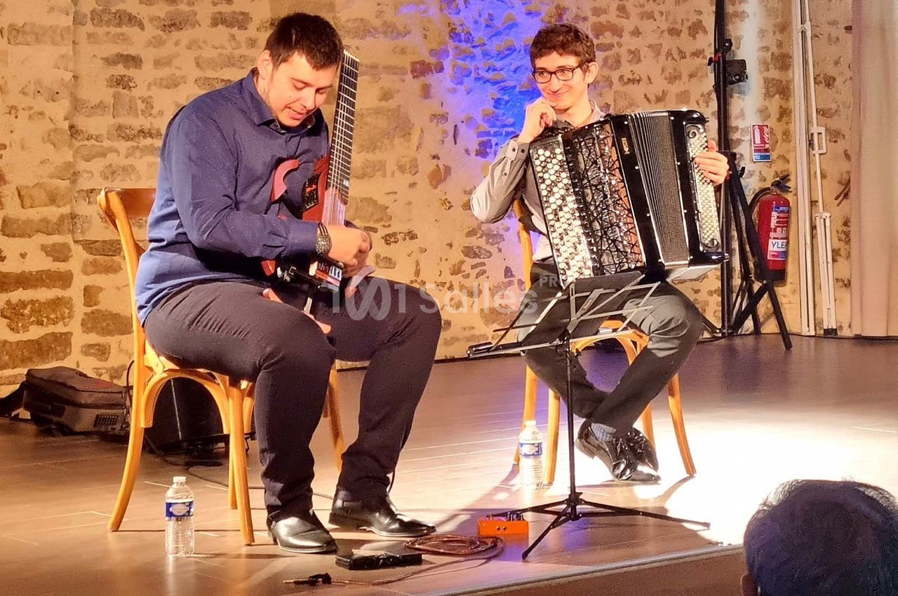 Deux musiciens assis jouent de la guitare électrique et de l'accordéon sur une scène éclairée, devant un mur en pierre.