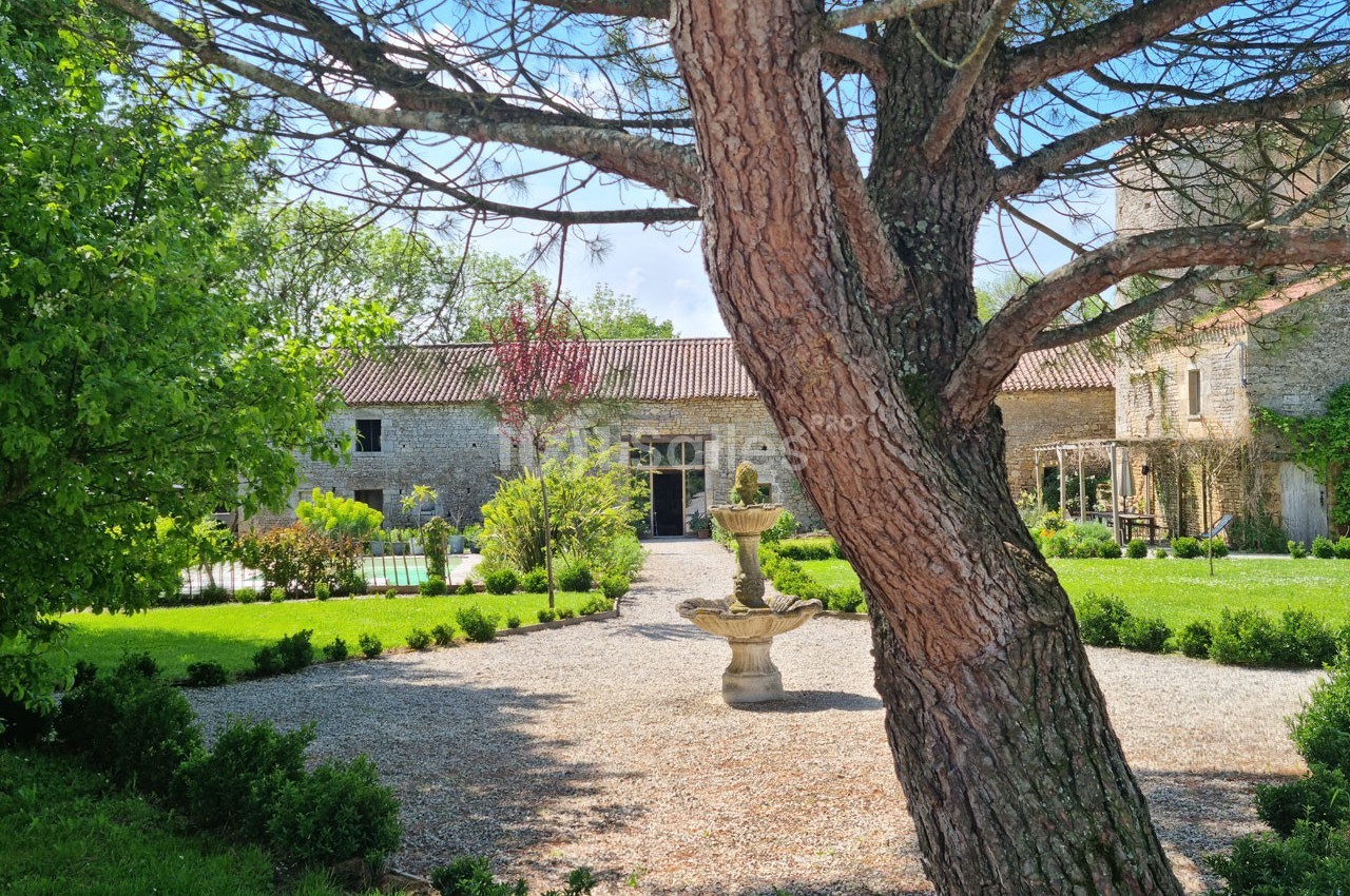 Cour d'une maison en pierre avec une fontaine centrale, entourée de verdure et d'arbres sous un ciel dégagé.