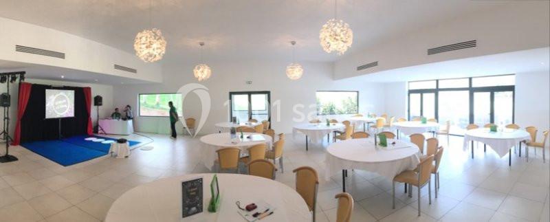 Location salle Marseille 11 (Bouches-du-Rhône) - La Bastide de la Salette #6 Salle lumineuse avec tables rondes dressées, chaises beige clair, projecteur et scène en arrière-plan.
