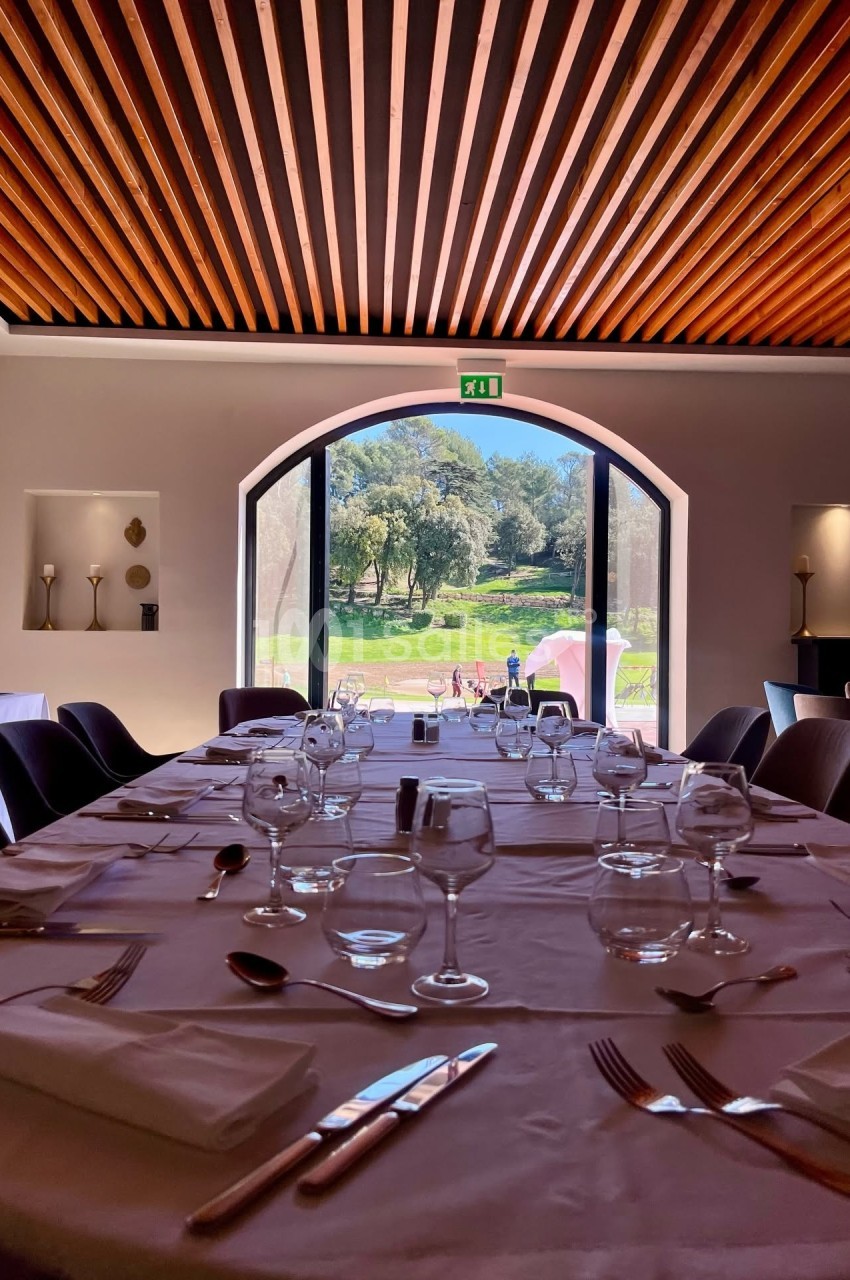 Salle de restaurant lumineuse avec table dressée, vue sur un jardin verdoyant à travers une grande baie vitrée.