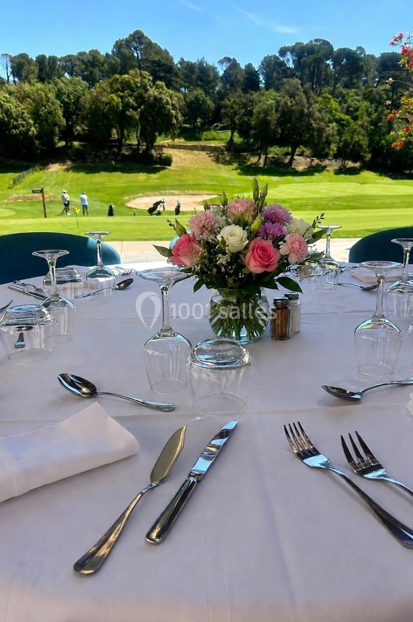 Location salle Marseille 11 (Bouches-du-Rhône) - La Bastide de la Salette #6 Table dressée avec nappes blanches, couverts et bouquet de fleurs, vue sur un terrain de golf en arrière-plan.
