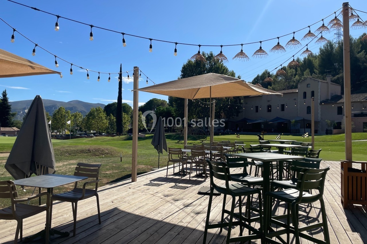 Location salle Marseille 11 (Bouches-du-Rhône) - La Bastide de la Salette #9 Terrasse en bois avec tables, chaises et parasols, éclairée par des guirlandes lumineuses, donnant sur un espace vert.