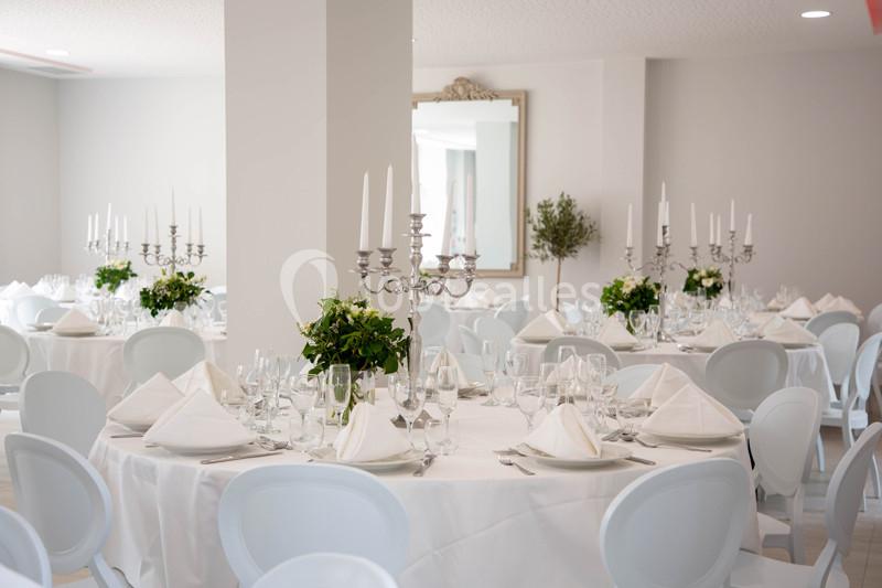 Salle de réception lumineuse avec tables rondes dressées, nappes blanches, chandeliers et décorations florales.