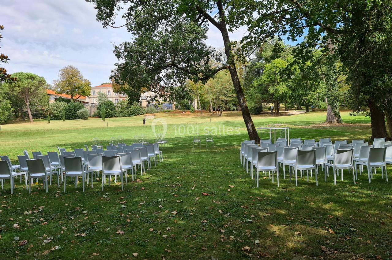 Chaises blanches disposées en extérieur sur une pelouse, entourées d'arbres, avec une arche au fond pour une cérémonie.