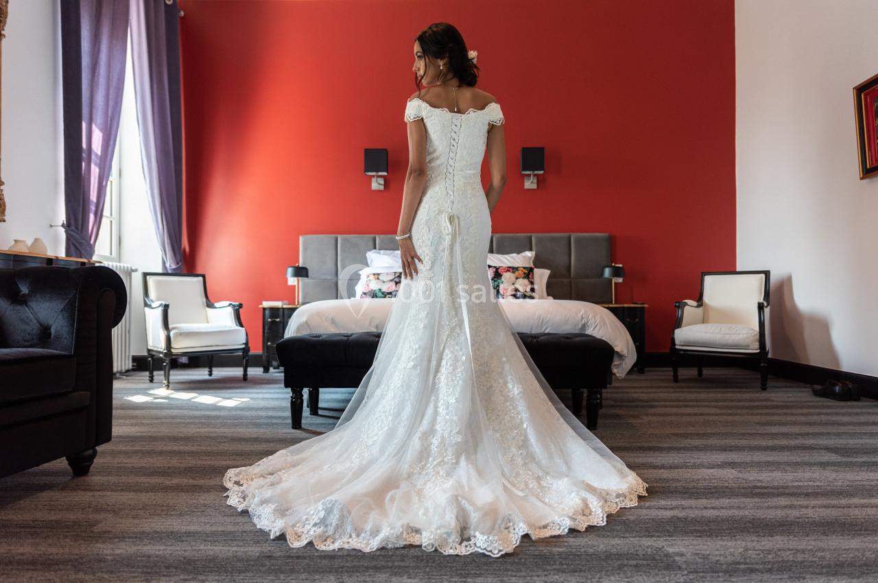 Une femme en robe de mariée blanche vue de dos dans une chambre avec un mur rouge et un lit au centre.