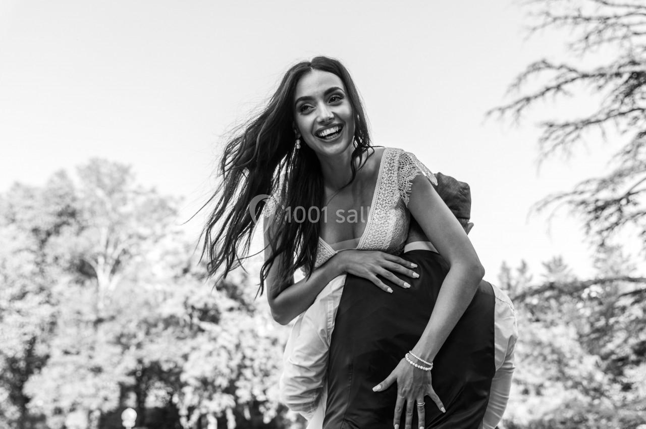 Femme souriante en robe blanche portée sur le dos d'une personne, dans un parc avec des arbres en arrière-plan.