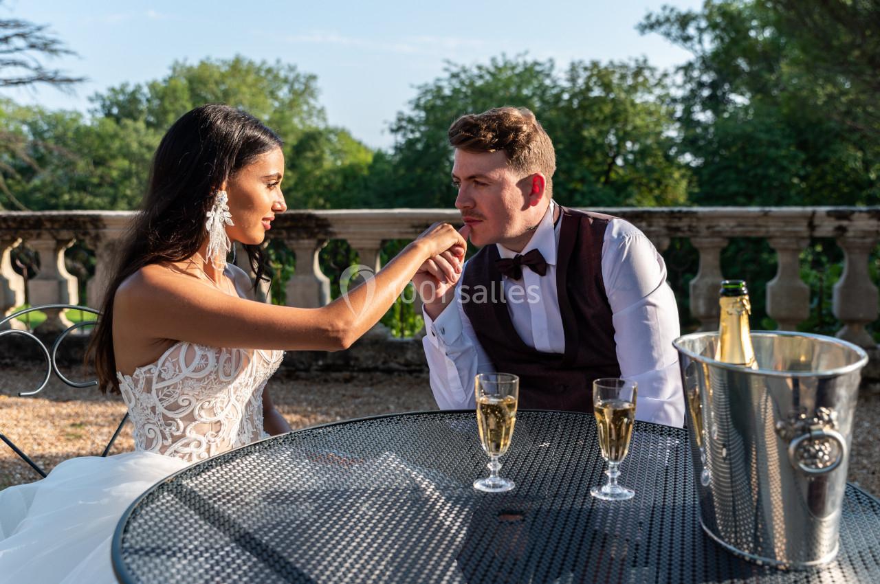 Une mariée et un marié assis à une table en extérieur, échangeant un geste tendre avec des coupes de champagne.