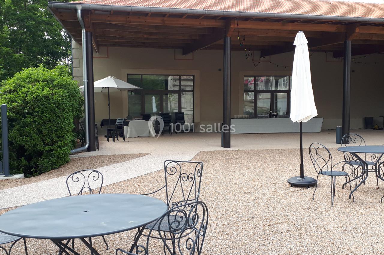Terrasse avec tables et chaises en métal noir, sol en gravier, parasols blancs et bâtiment avec toit en tuiles.