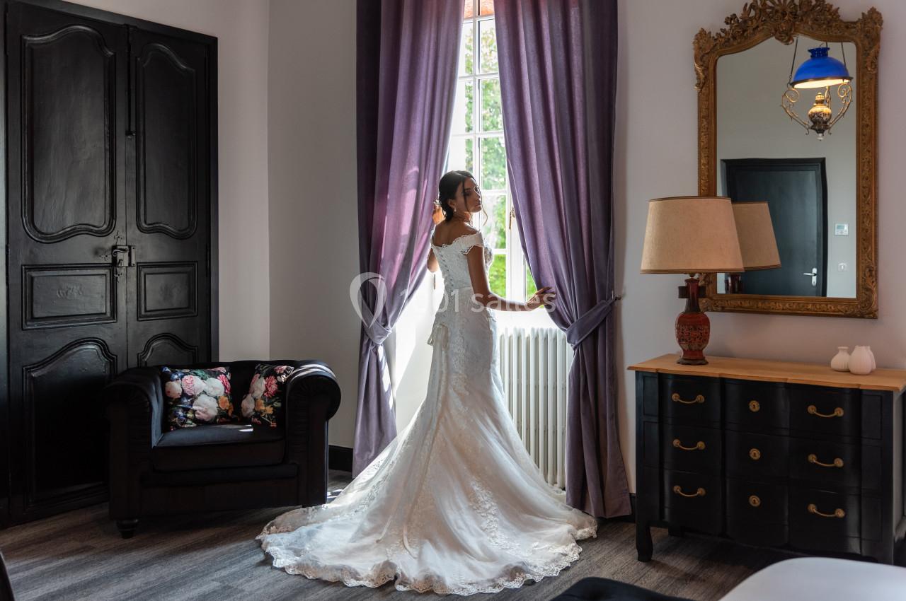 Une femme en robe de mariée regarde par une fenêtre dans une pièce élégante avec des rideaux violets et un miroir doré.
