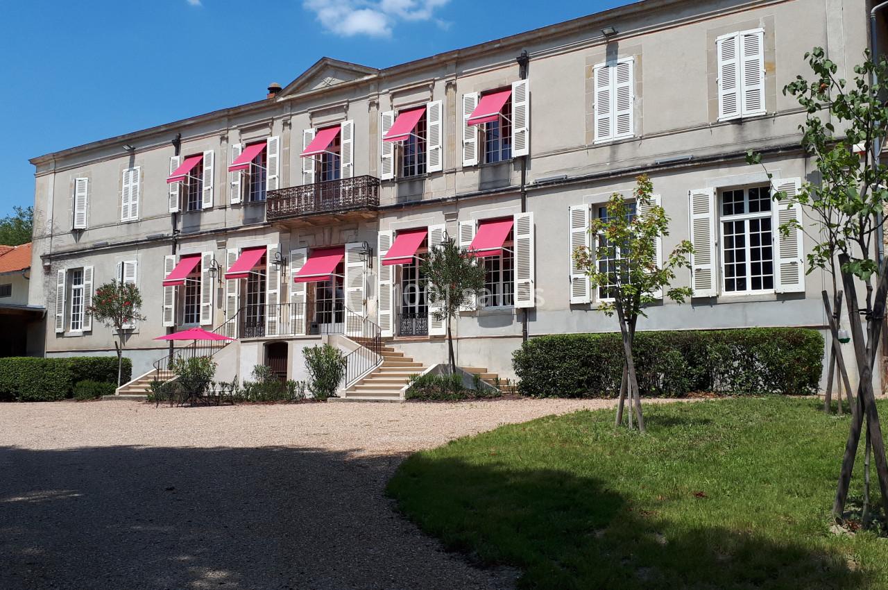 Façade d'un grand bâtiment ancien avec volets blancs et stores rouges, entouré de verdure et d'une allée gravillonnée.