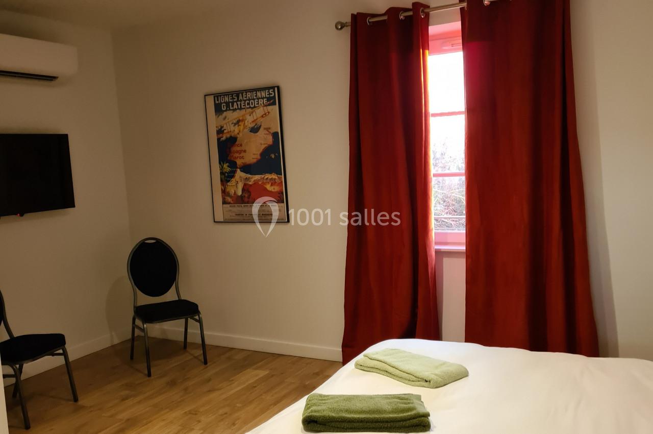 Chambre lumineuse avec lit double, draps blancs, serviettes vertes, rideaux rouges et télévision murale.