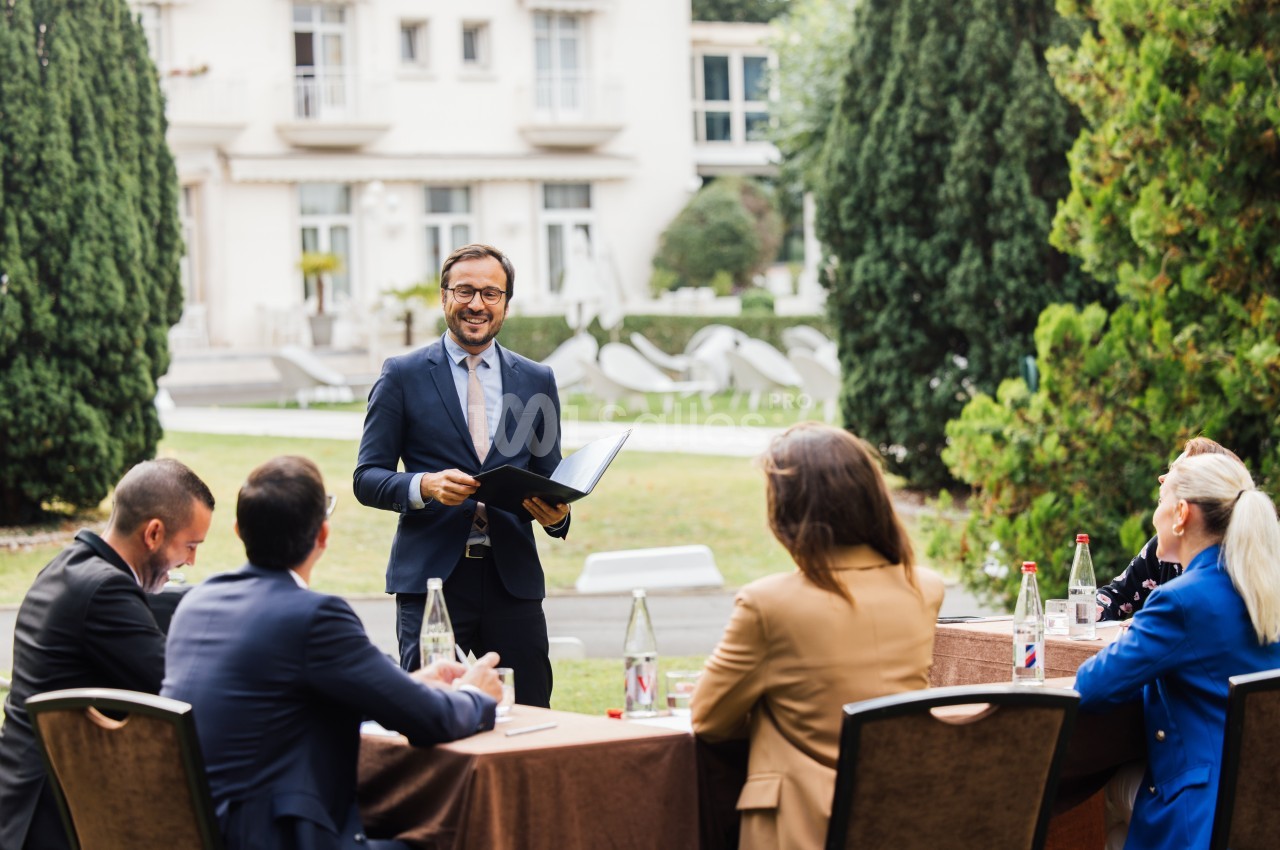 Un homme en costume présente un document à un groupe de personnes assises autour d'une table en extérieur.