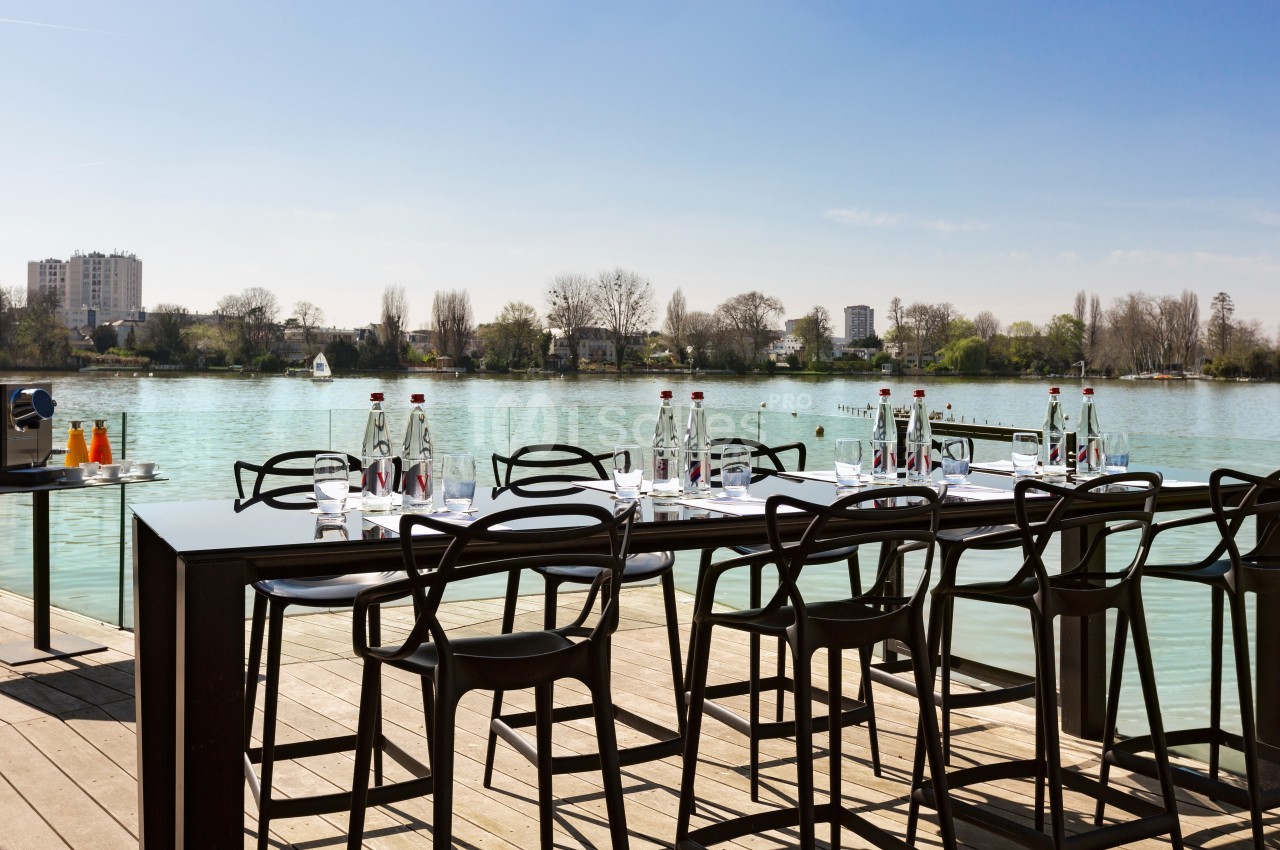Table dressée avec chaises modernes sur une terrasse en bois, vue sur un lac et des bâtiments en arrière-plan.