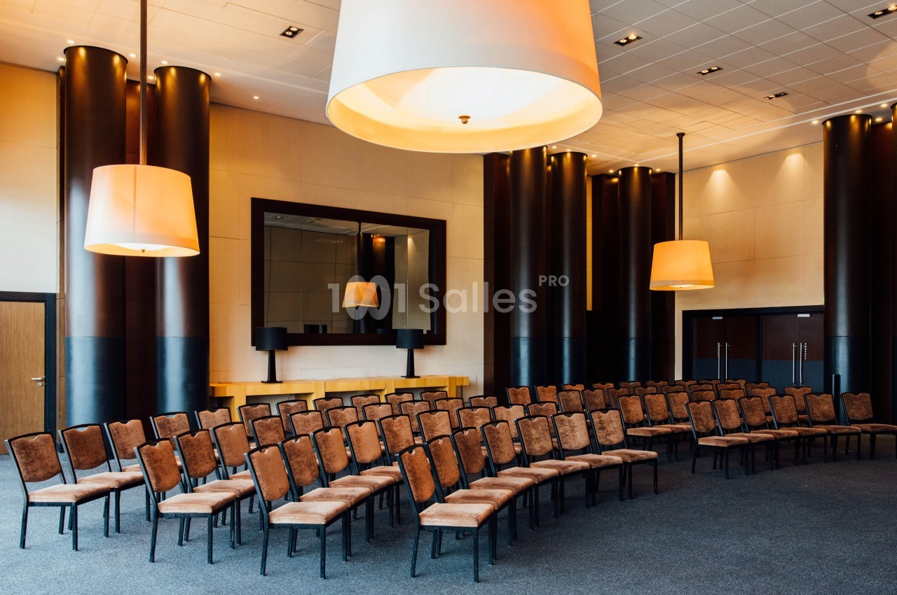 Salle de conférence avec des rangées de chaises vides, éclairée par des suspensions lumineuses modernes.
