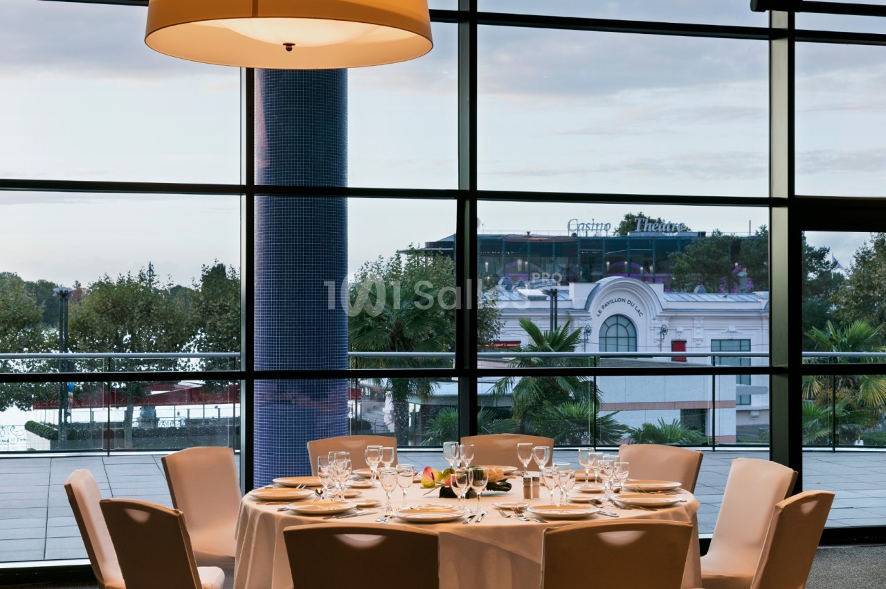 Table dressée avec vaisselle élégante devant une grande baie vitrée offrant une vue sur un bâtiment et des palmiers.
