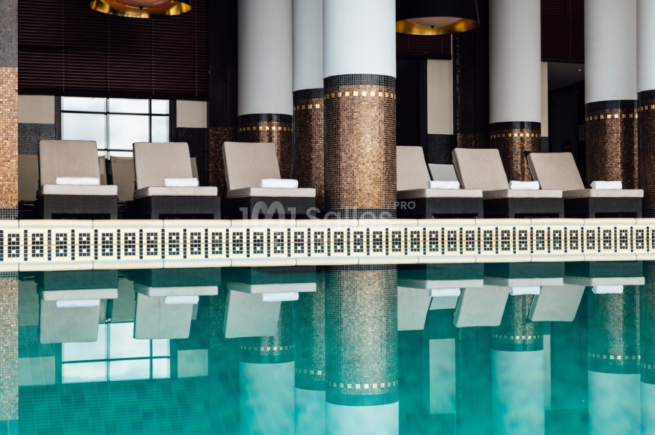 Chaises longues alignées près d'une piscine intérieure, entourée de colonnes et de murs décorés de mosaïques.