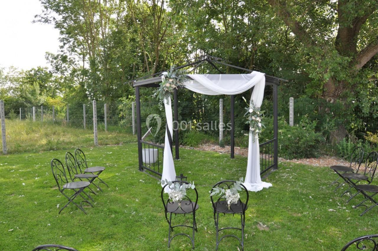 Pergola décorée de voilages blancs et de fleurs, entourée de chaises noires sur une pelouse verdoyante.