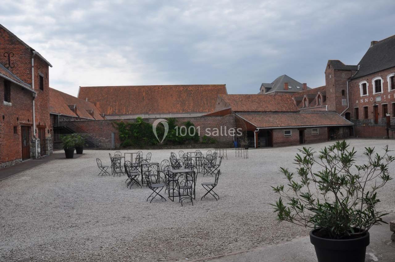 Cour pavée avec des tables et chaises en fer forgé, entourée de bâtiments en briques sous un ciel nuageux.