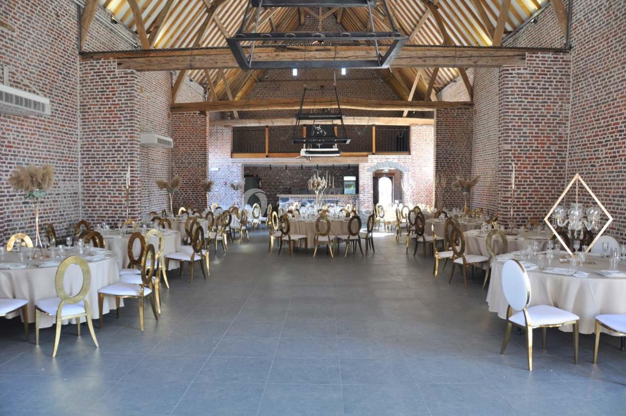 Salle de réception spacieuse avec murs en briques, poutres apparentes et tables rondes dressées pour un événement.