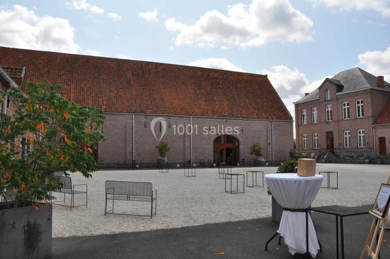 Cour pavée avec une grande bâtisse en briques et toit en tuiles, entourée de bancs et d'arbres en pot.