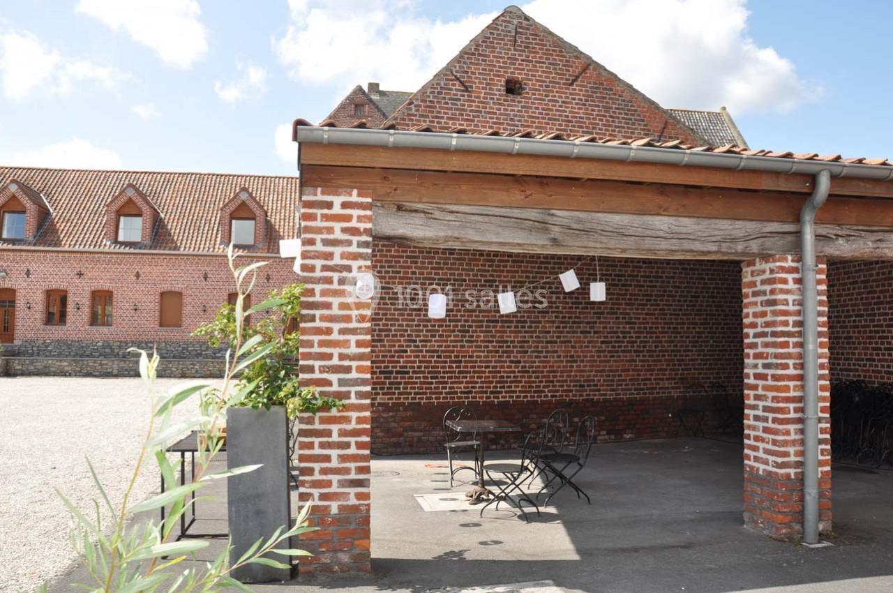 Abri en briques avec toit en bois, tables et chaises noires, situé dans une cour pavée devant un bâtiment en briques.