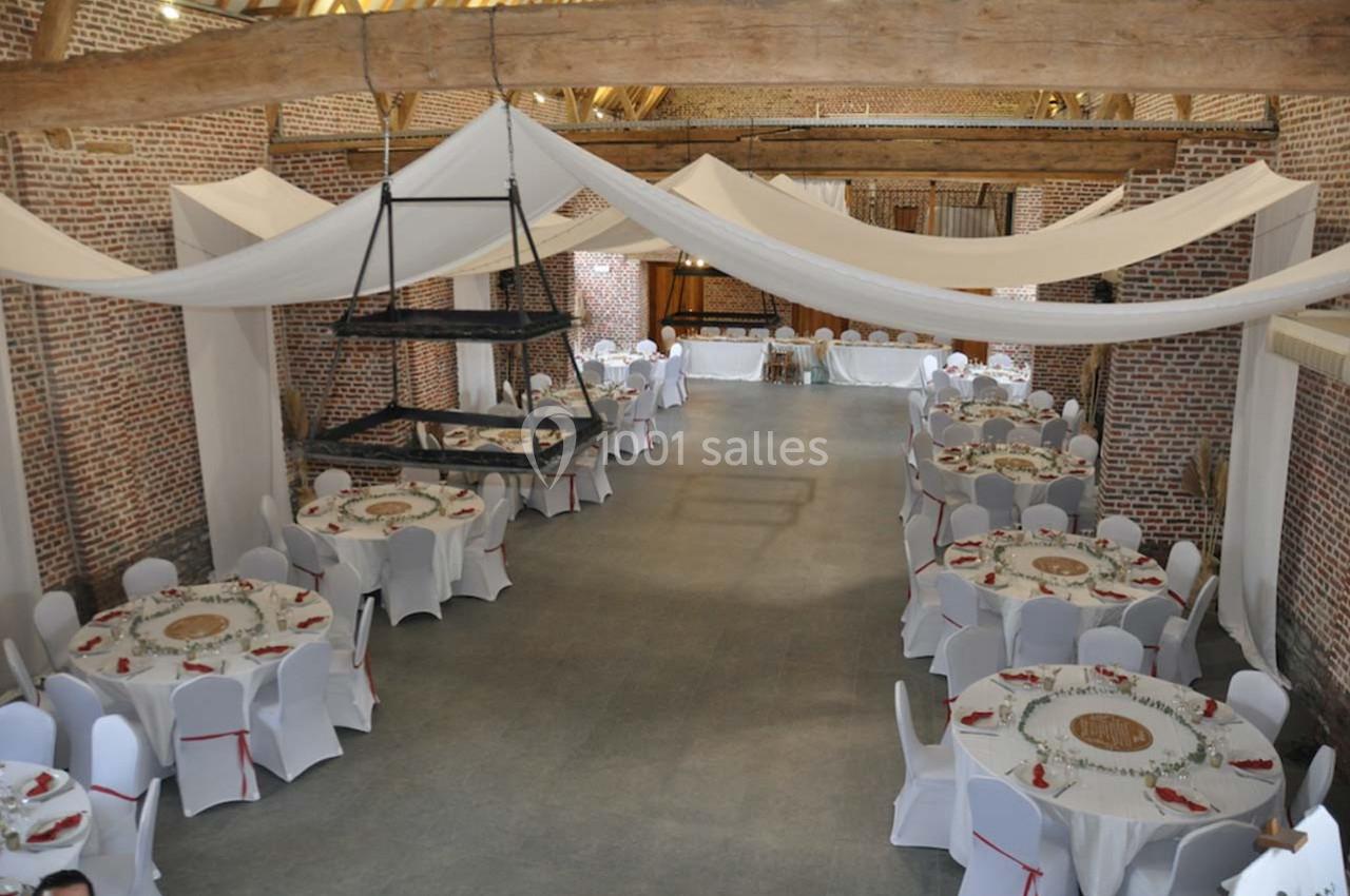 Salle de réception avec murs en briques, poutres apparentes, tables rondes dressées et décorations en tissu suspendues.
