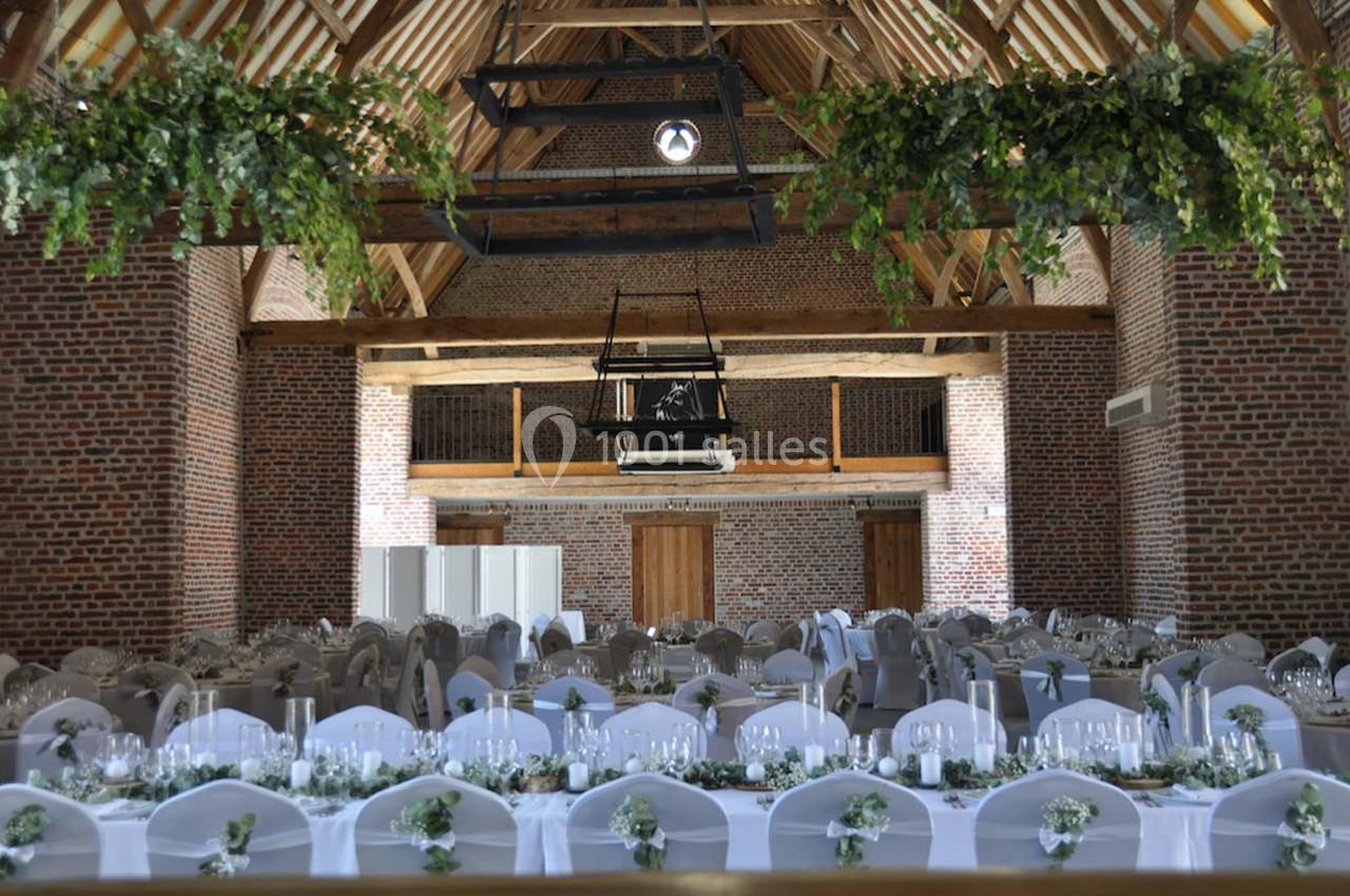 Salle de réception avec des tables décorées de nappes blanches et de compositions florales, sous une charpente en bois.