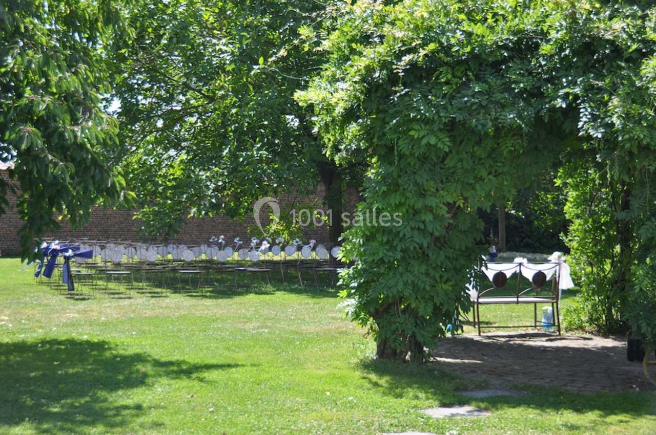 Un jardin verdoyant avec des tables dressées sous les arbres et une tonnelle couverte de feuillage.
