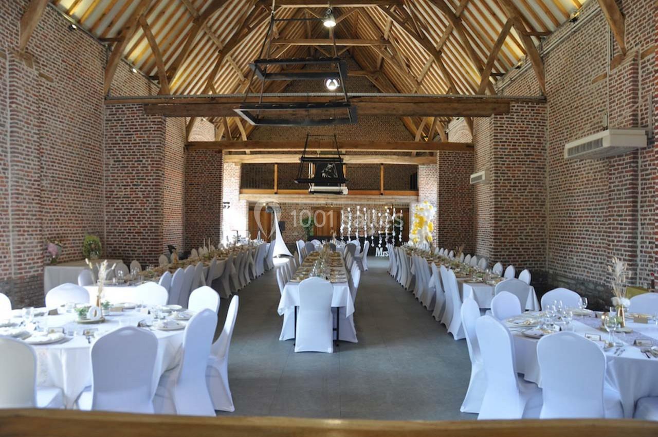 Salle de réception en briques avec tables rondes dressées et chaises blanches, sous une charpente en bois apparente.