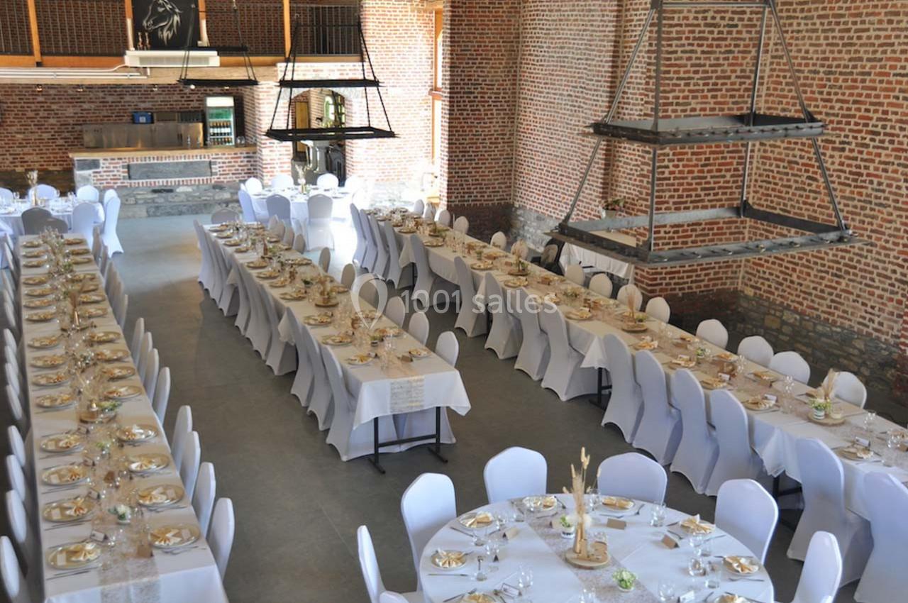 Salle de réception avec de longues tables dressées, nappes blanches et chaises couvertes, dans un décor en briques…