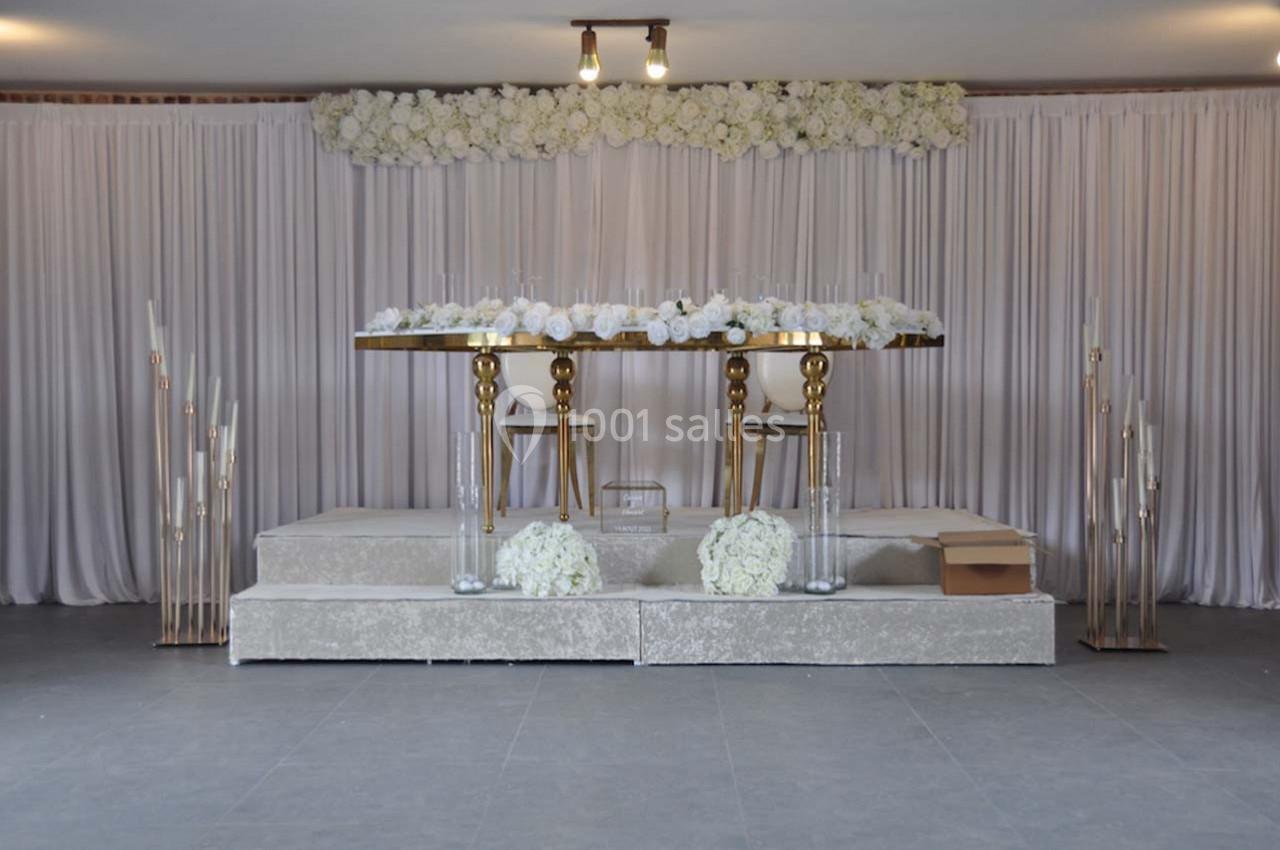 Table décorée avec des fleurs blanches sur une estrade, entourée de rideaux clairs et de chandeliers modernes.