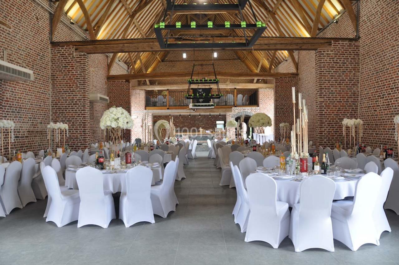 Salle de réception avec tables rondes dressées, chaises blanches et décoration florale, dans un cadre en briques et poutres…