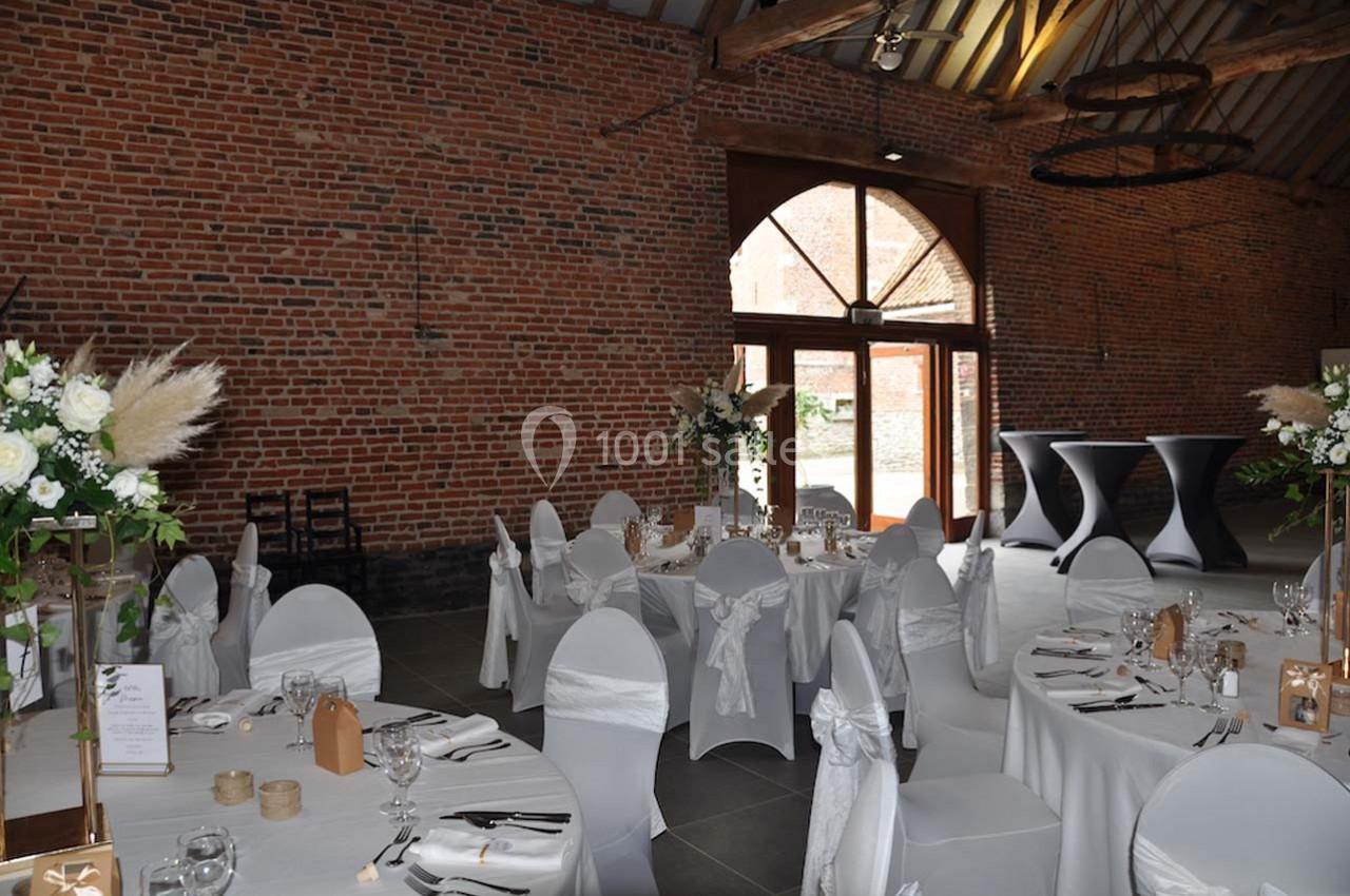 Salle de réception avec murs en briques, tables rondes dressées, chaises blanches et grandes ouvertures vitrées.