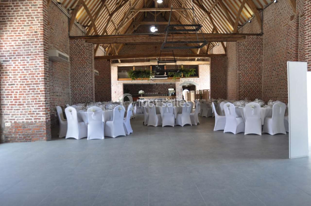 Salle de réception en briques avec poutres apparentes, tables rondes dressées et chaises blanches houssées.
