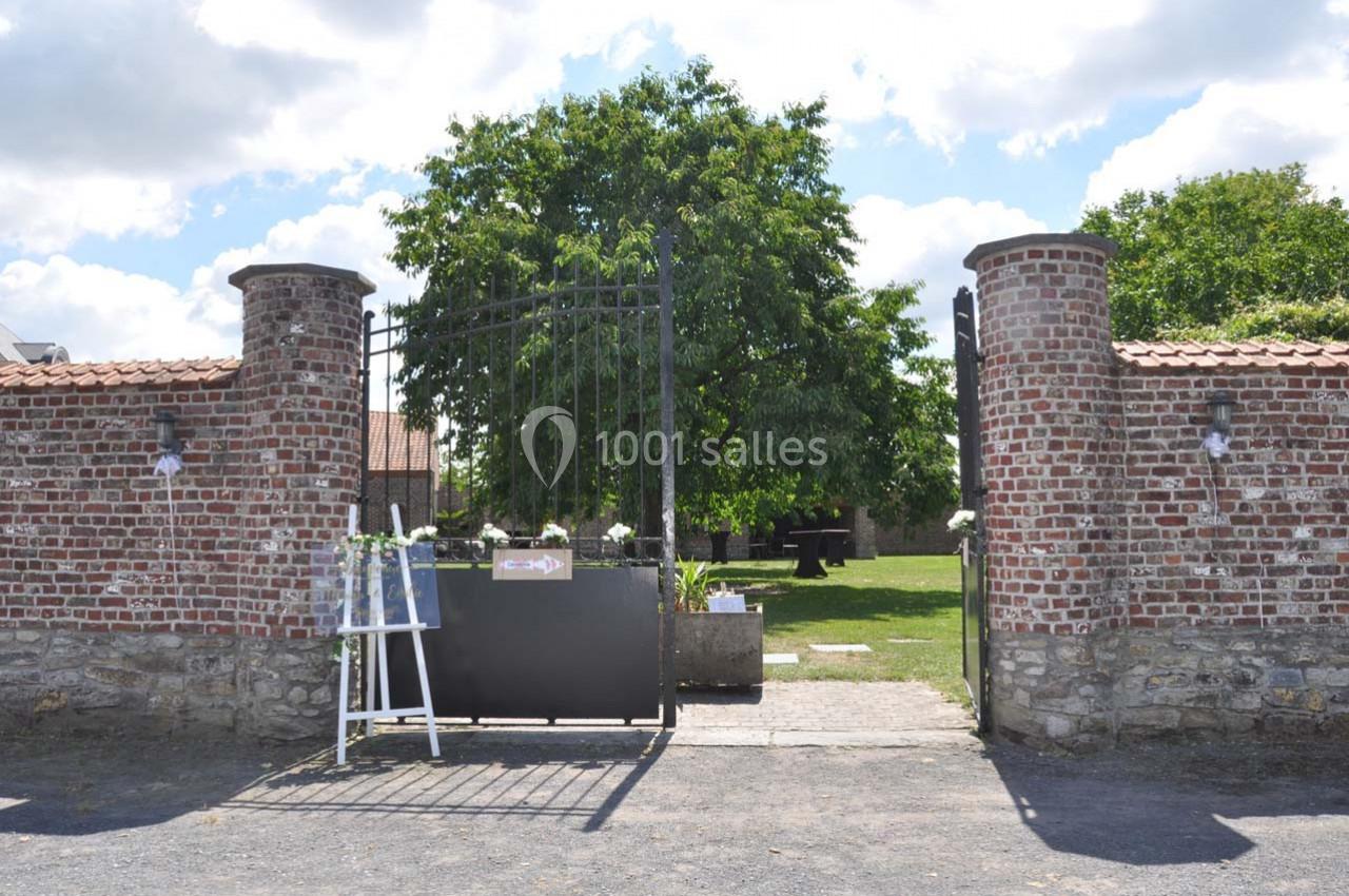 Entrée d'une cour avec portail en fer forgé, murs en briques et un arbre au centre sous un ciel dégagé.
