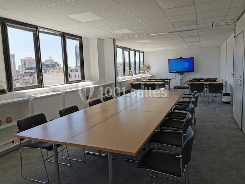 Salle de réunion lumineuse avec grande table, chaises noires, étagères, écran mural et vue sur des immeubles urbains.