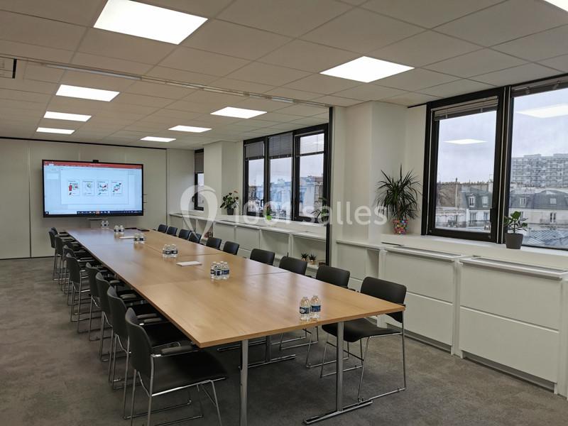 Salle de réunion lumineuse avec une grande table rectangulaire, des chaises noires, un écran et des fenêtres donnant sur la…