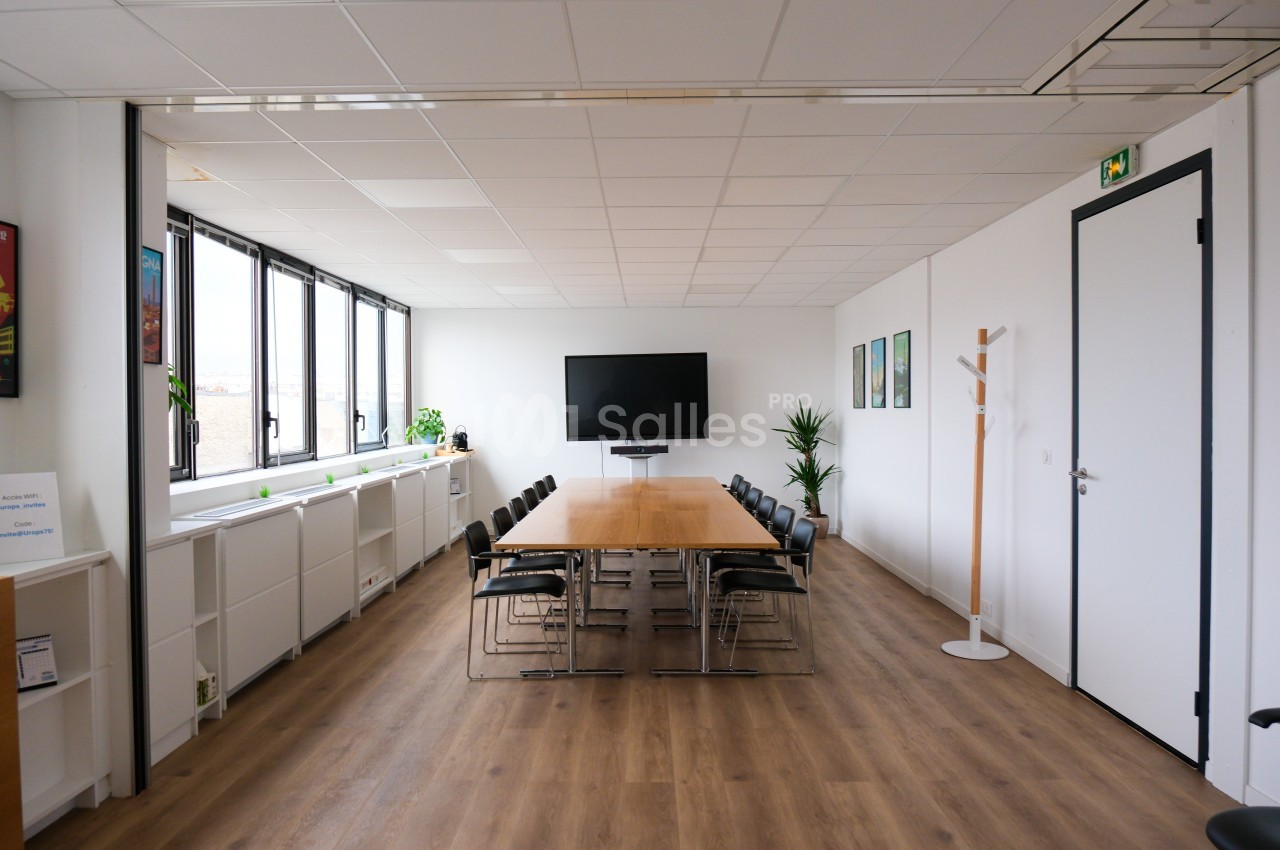 Salle de réunion lumineuse avec une grande table en bois, des chaises noires, un écran mural et des plantes décoratives.