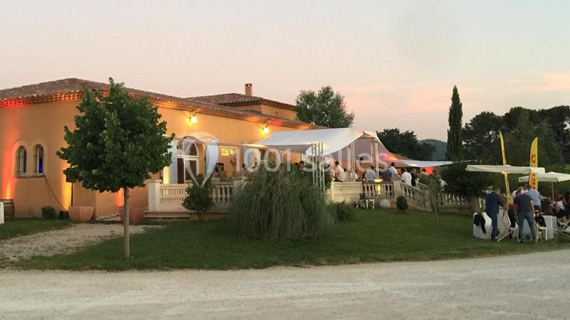 Bâtiment de style méditerranéen avec terrasse éclairée au crépuscule, entouré de verdure et d'invités.