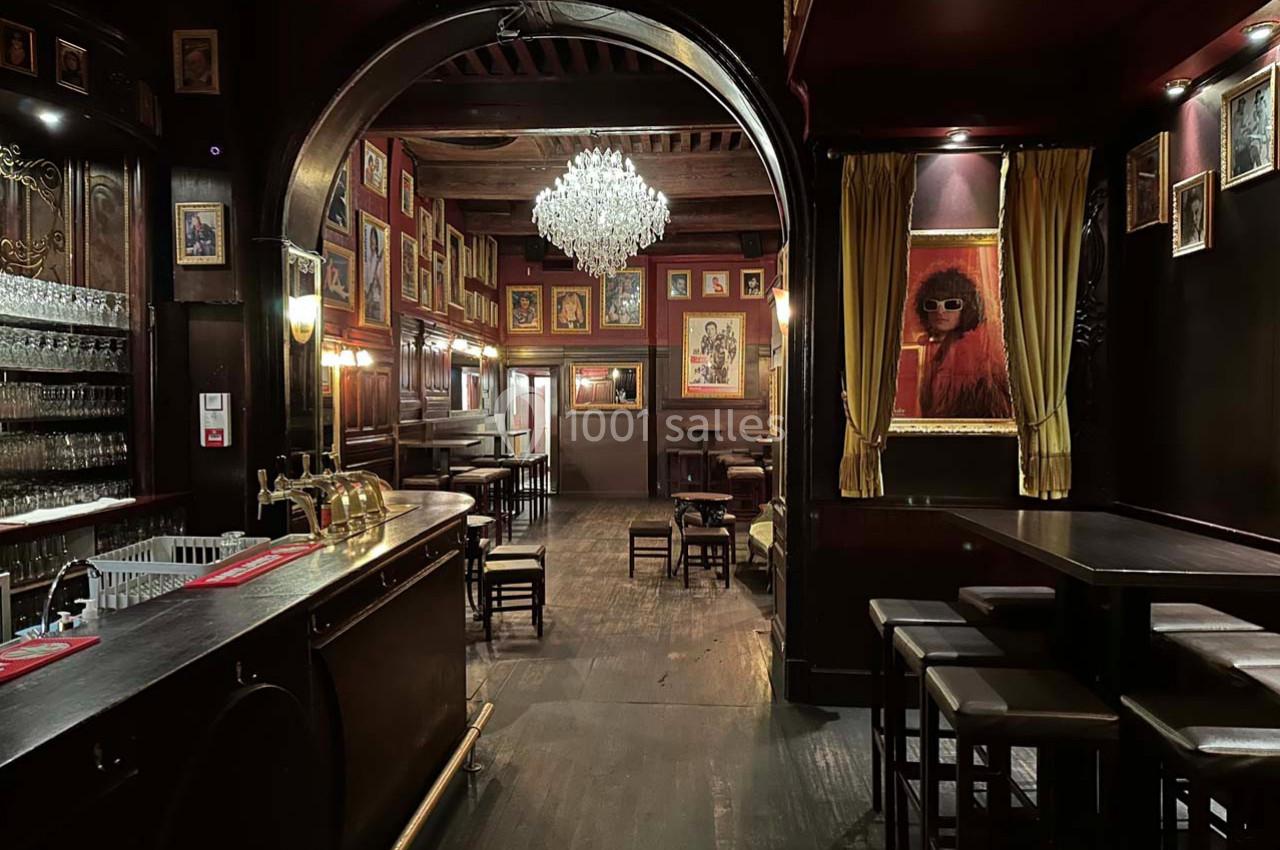 Intérieur d'un bar chaleureux avec comptoir en bois, lustre central, tableaux encadrés et ambiance tamisée.