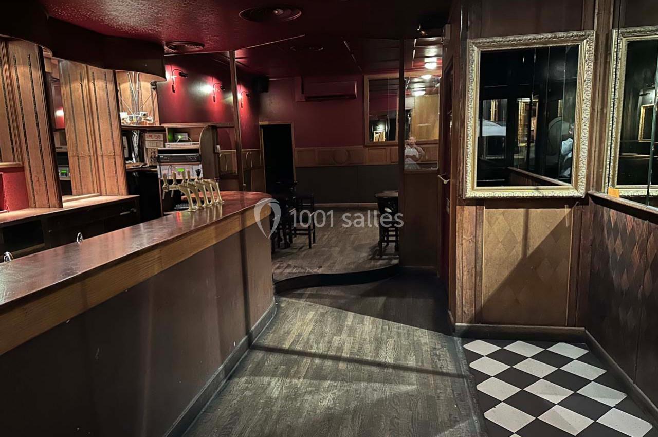 Intérieur d'un bar sombre avec comptoir en bois, sol en damier près de l'entrée et quelques tables au fond.