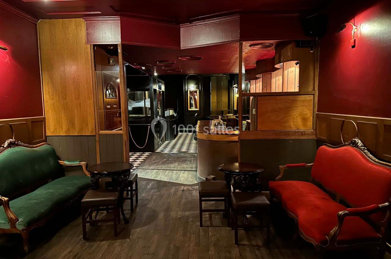 Intérieur d'un salon avec fauteuils rouges et verts, tables basses en bois, éclairage tamisé et sol en parquet.