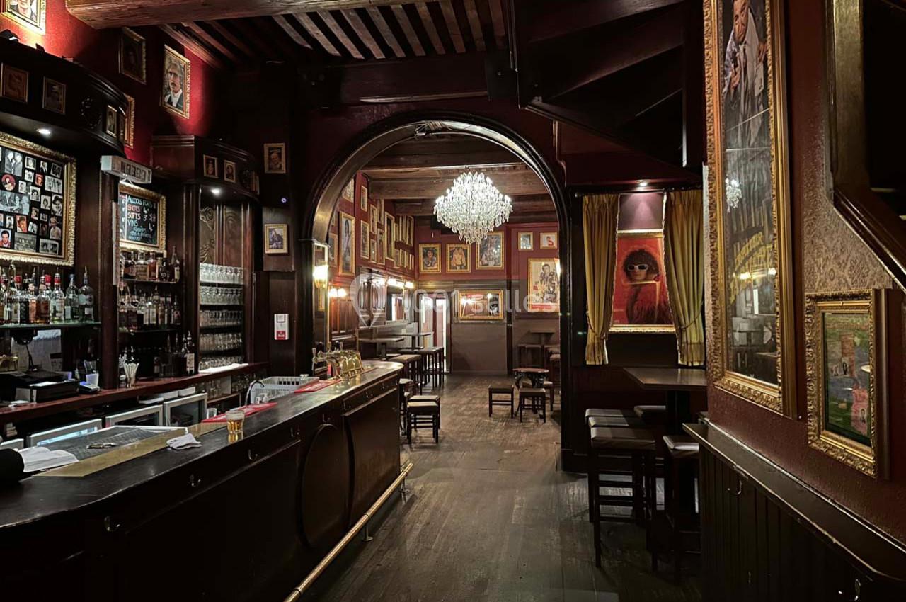 Intérieur d'un bar chaleureux avec un comptoir en bois, des murs décorés de cadres et un lustre central.
