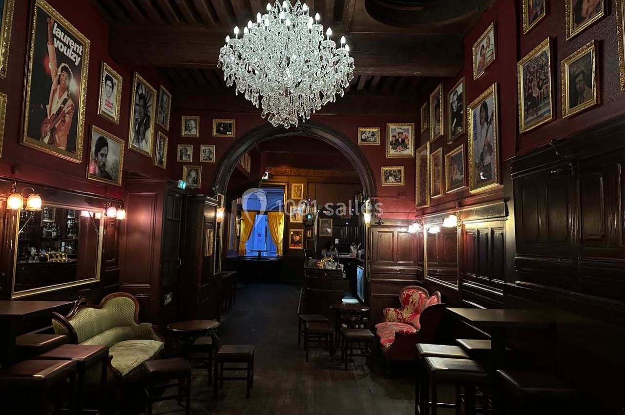 Intérieur d'un bar à l'ambiance vintage, avec un lustre central, des fauteuils anciens et des murs ornés de cadres.