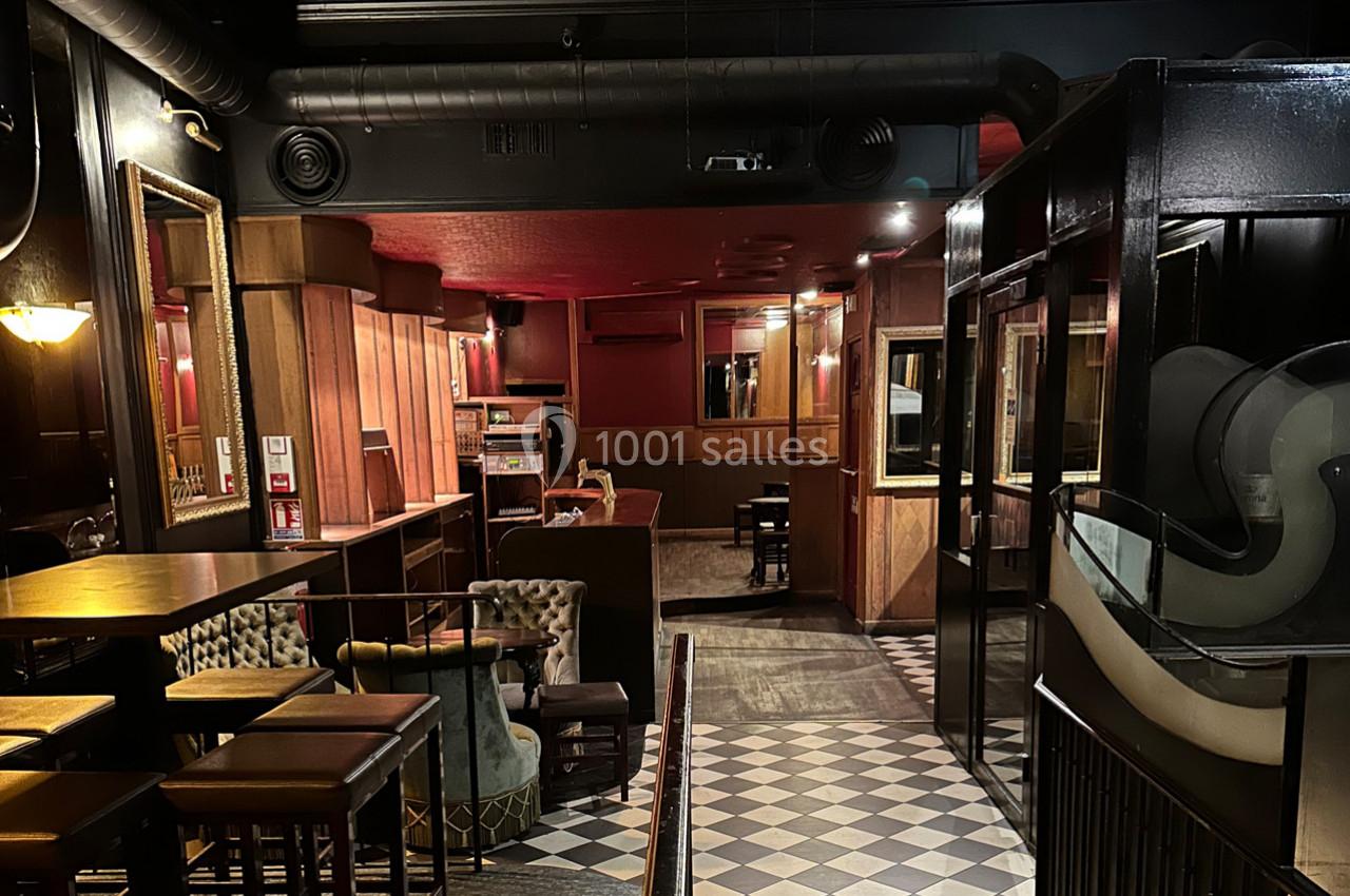 Intérieur d'un bar avec mobilier en bois, fauteuils, sol à carreaux et éclairage tamisé.
