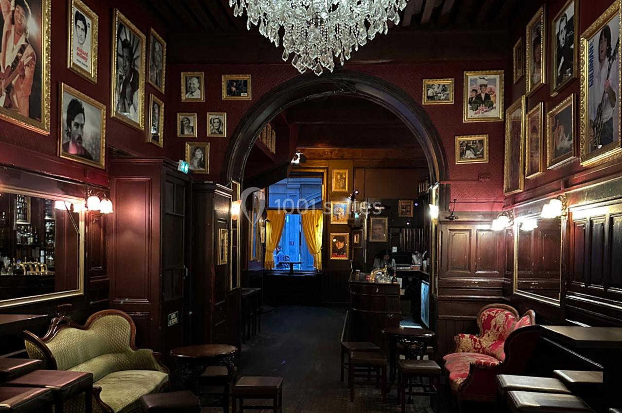 Intérieur d'un bar ou salon avec murs rouges ornés de photos encadrées, mobilier ancien et lustre en cristal.